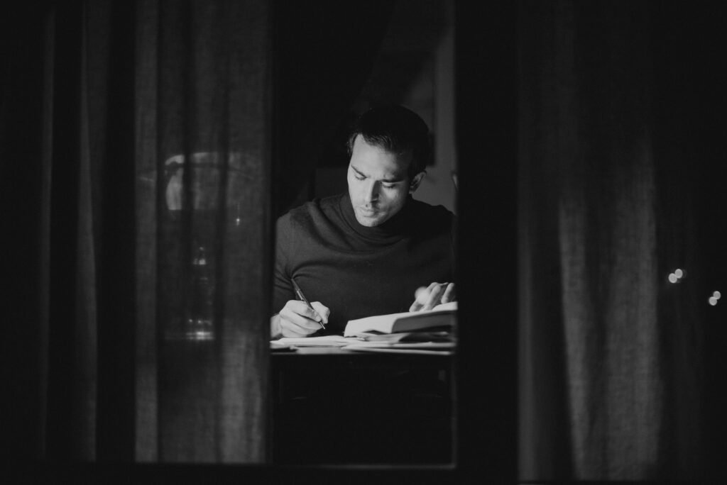 A man deeply focused on studying at night, captured in black and white from an indoor perspective.